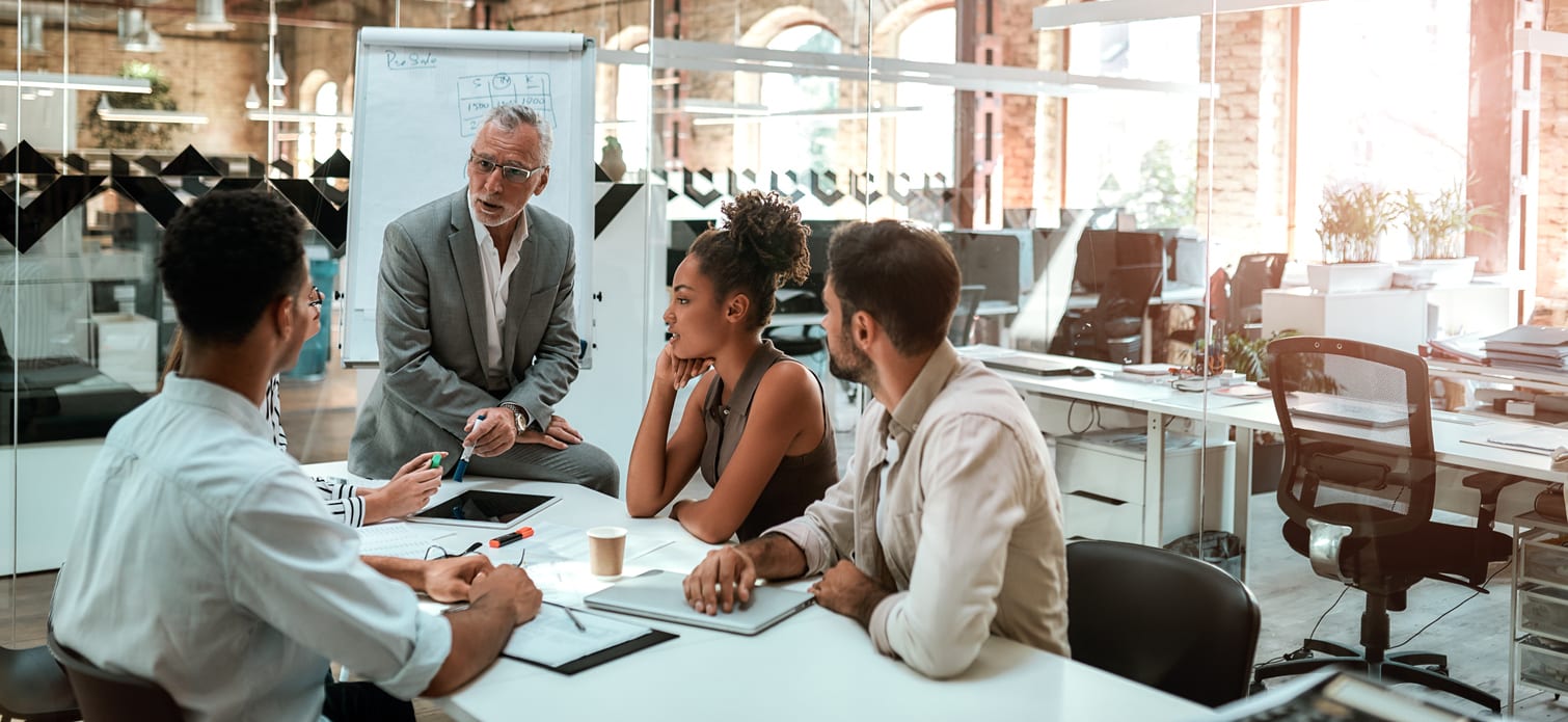 Mature businessman talking about something with his young colleagues while sitting together at the office table in the modern office.