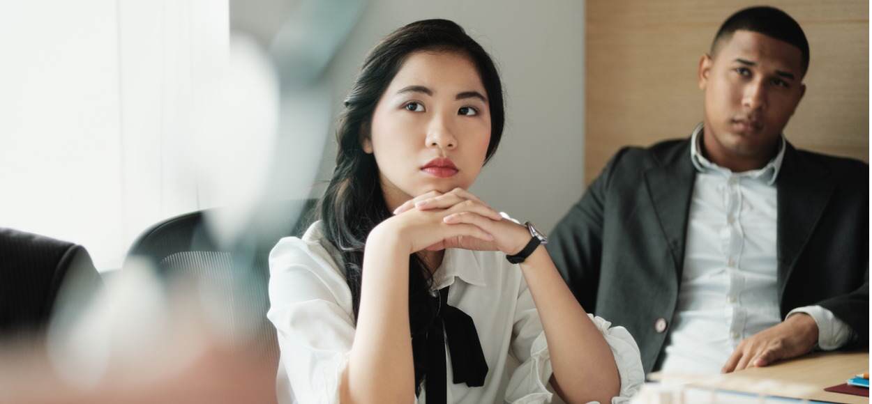 Young woman crosses hands as male colleague sits behind her looking stern