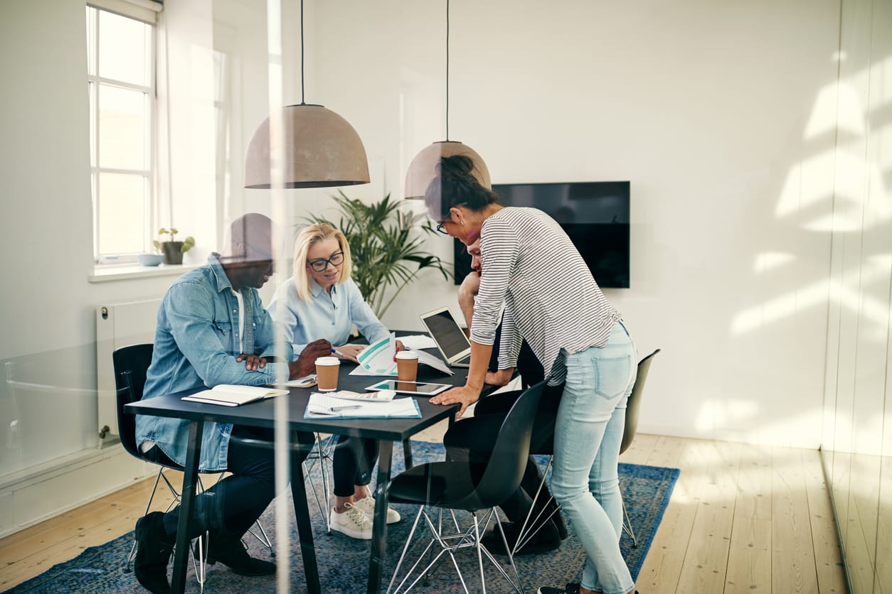 Diverse group of young businesspeople sitting together around a table inside of a glass walled office discussing paperwork