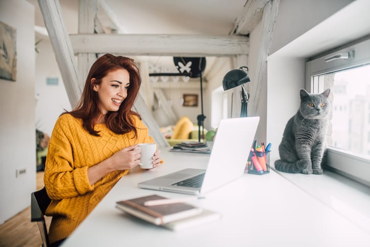 Smiling young redhead holding a cup of coffee while working on her laptop in her home office with a cat sitting in a window