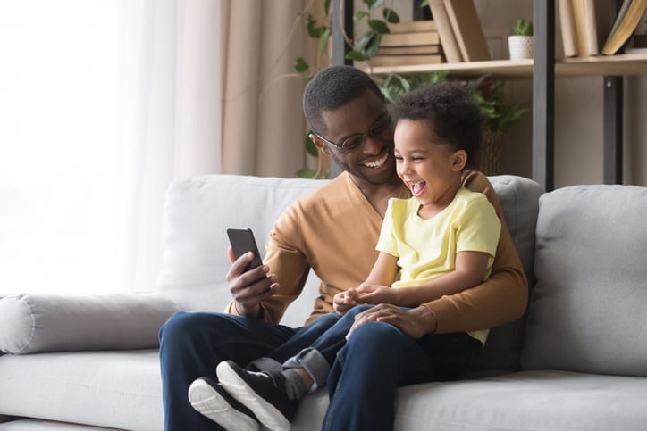 Happy african dad and toddler son laughing looking at cellphone