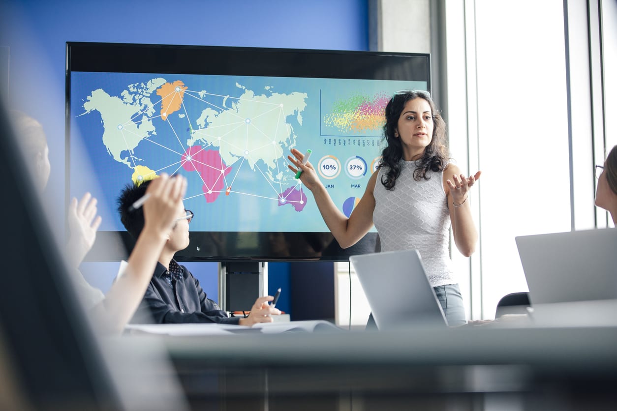 A mixed race woman is giving presentation. This confident individual stands in front of a data display monitor, which has colourful world graphics displayed on it. Image focus technique, layers and point of view, help the image to be realistic, as if the viewer is spying on the participants. This lady is a natural leader, she is answering questions from her colleagues. The colour blue is strong in this image.