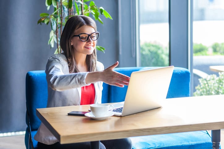 Portrait of happy beautiful stylish young woman in glasses sitting, looking at her laptop screen on video call and giving hand to handshake, toothy smile. indoor studio shot, cafe, office background.