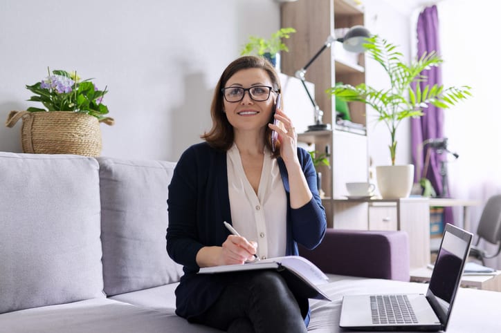 Mature woman working remotely at home, business female freelancer sitting on sofa with laptop, talking on phone, taking notes in business notebook.