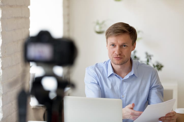 Serious confident young professional lecturer coach mentor speaker sitting at desk with computer in front of camera, recording educational lecture for personal channel, presenting financial report.