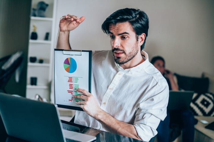 Businessman talking to his colleagues about business plan in video conference. Young man working at home. Businessman discussing graph with colleagues via video call. Businessman holding financial graph against his laptop. Handsome young man having video call at home. Shot of a young man using a laptop for web conference.