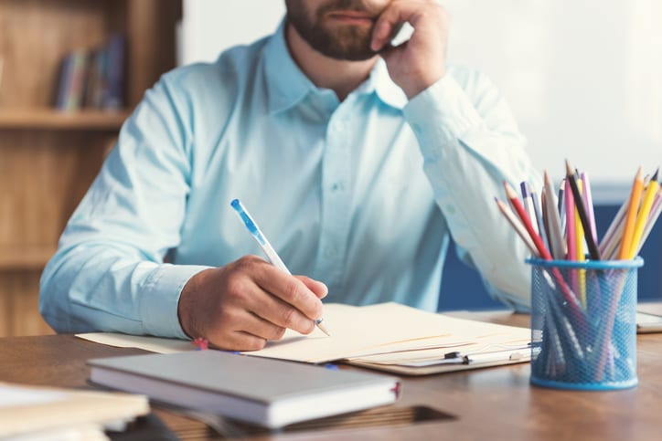 Close up of hands and body of bearded man sitting at table and writing some task by pen on prepare. He is leaning his cheek on one hand