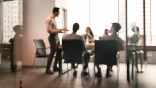 Business Presentation, Blurred Background. Businessman Giving Speech During Seminar With Coworkers In Office, Standing At Desk In Boardroom, Diverse People Sitting At Table And Listening To Speaker