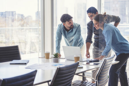 Business team working on a laptop computer. Three people are wearing casual clothing. They are standing in a board room. Multi ethnic group with Caucasian and Latino men and women. They are all happy and smiling