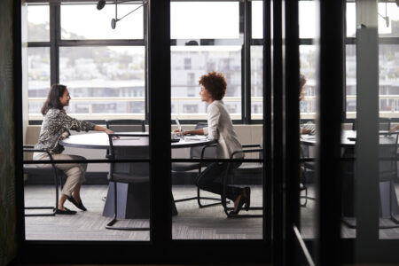 Two millennial businesswomen meeting for a job interview, full length, seen through glass wall