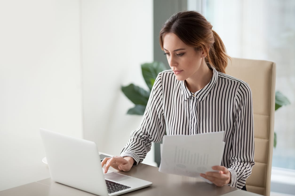 Serious focused businesswoman typing on laptop holding papers preparing report analyzing work results, female executive doing paperwork at workplace using computer online software for data analysis