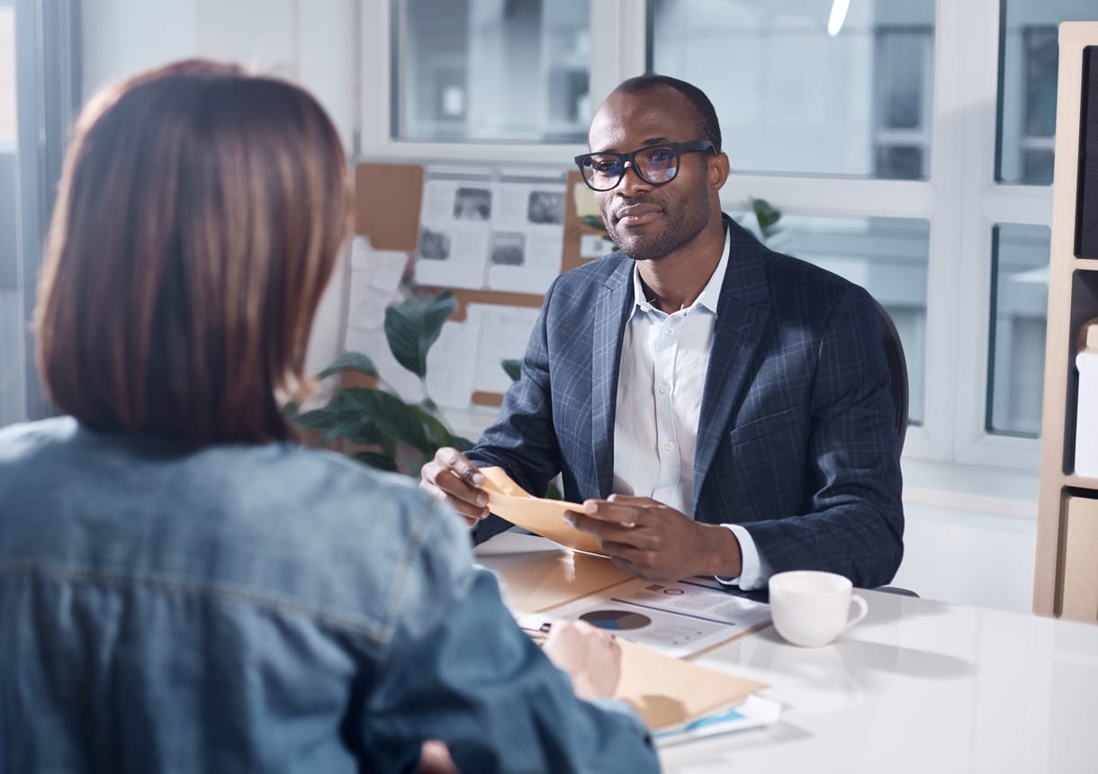 Listen attentively. Skillful young pleasant african businessman in suit is sitting at table and holding papers while looking at responsible colleague female who sitting in front of him