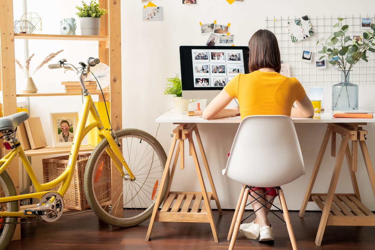 Rear view of young brunette female sitting on white chair in front of computer monitor while looking throguh collection of photos