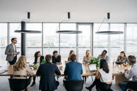 A photo of a business meeting in a modern office with large windows. A businessman is standing up while his colleagues are sitting down. They are smartly dressed. Horizontal daylight indoor photo.