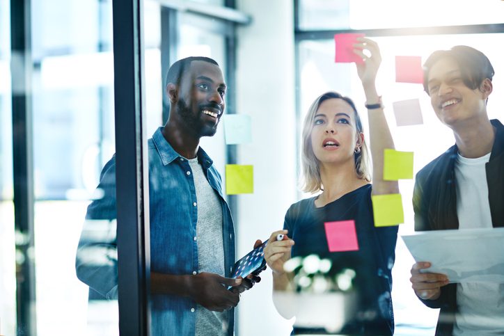 Two male employees and a female employee putting sticky notes on a glass wall whilst holding tablets and paper