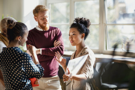 Colleagues standing in a small group discussing something. One of the women is holding documents and gesturing with her hands as the others watch and listen.