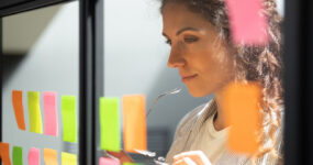 Head shot close up thoughtful young female boss team leader looking at kanban scrum glass window board, developing start up project marketing strategy, managing workflow, checking tasks at office.