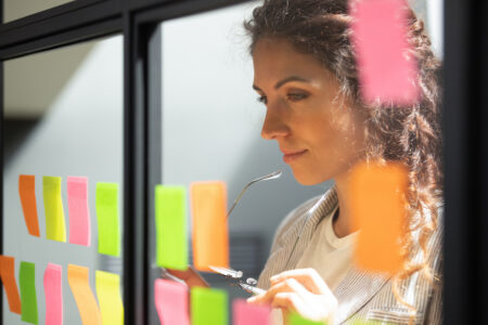 Head shot close up thoughtful young female boss team leader looking at kanban scrum glass window board, developing start up project marketing strategy, managing workflow, checking tasks at office.