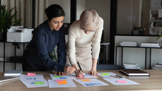 Multiracial businesswomen work on project statistics, prepare financial report use post-it memo notepapers making notes do sales overview, colleagues analysing charts data, teamwork brainstorm concept