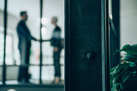 Business people shaking hands in the office. Blurred shot of two business persons shaking hands in a modern office. Business partners shaking hands, seen through glass wall in the office. Defocused shot of a businessman and businesswoman handshaking. The view is through glass.