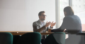 Focused confident young businessman in eyewear explaining project details to concentrated male client. Serious hr manager listening to skilled job applicant self presentation at interview in office.