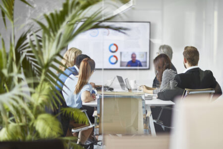 Rear view photographed through window of diverse male and female executive team video conferencing with CEO in office board room.