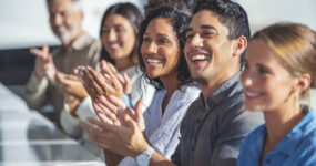 Group of business people applauding a presentation. They are sitting at a table in a sunny room. There are several ethnicities present including Caucasian, African and Latino