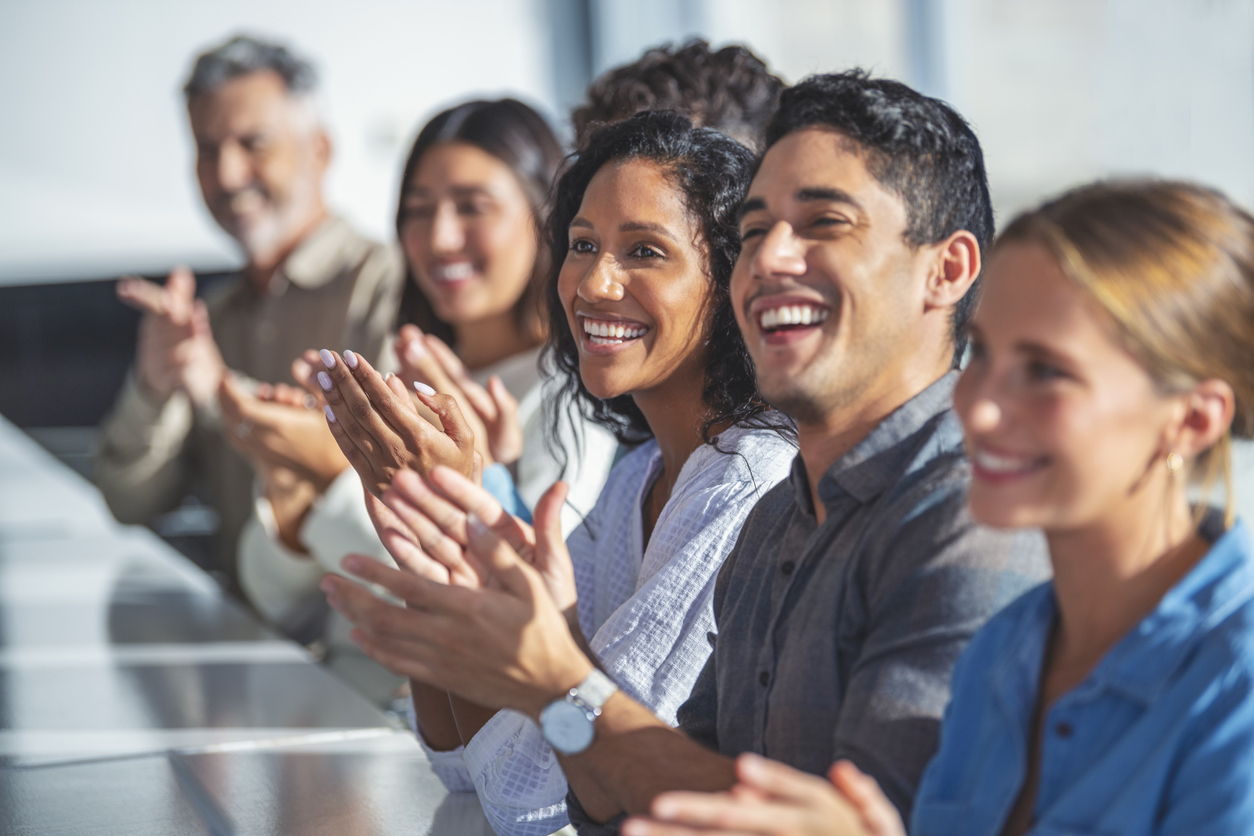 Group of business people applauding a presentation. They are sitting at a table in a sunny room. There are several ethnicities present including Caucasian, African and Latino