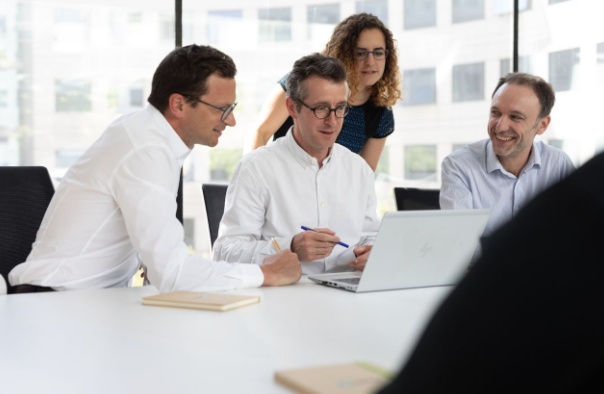 three men and one women looking at a single laptop whilst in a meeting