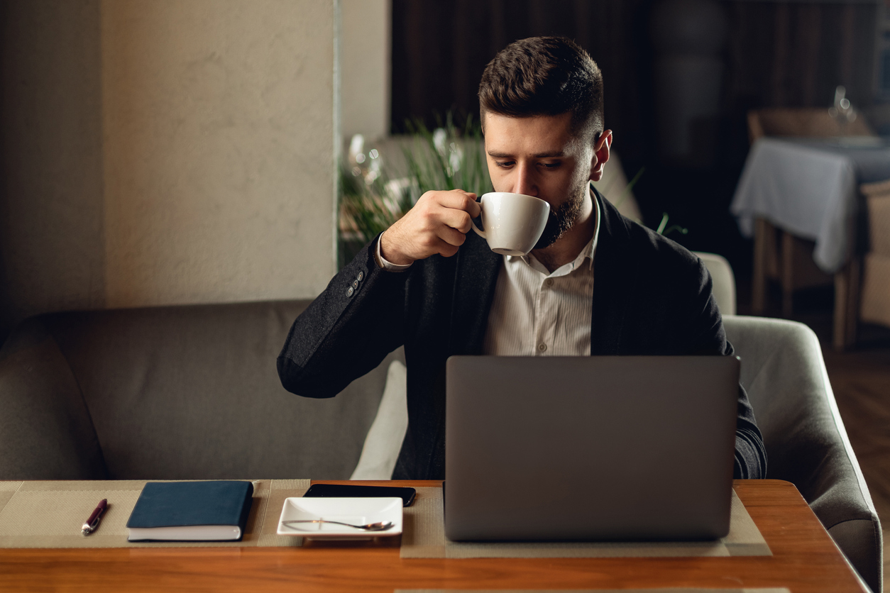 Man in black jacket drinking from a mug whilst working on a laptop