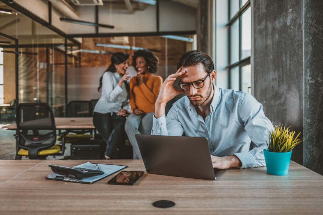 Lonely and sad businessman at office of company. Photo of Sad businessman with female team in the background. Male office manager unable to cope with too many tasks from his boss and supervisor, suffering stress, pressing her head with desperate expression.