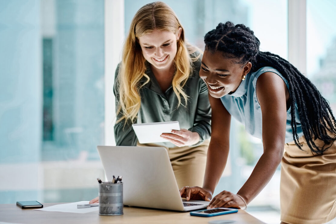 Two females leaning over laptop screen smiling and discussing work